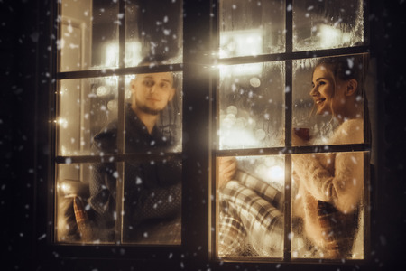 Young romantic couple is spending time together at home in pajamas.Sitting on window sill with garland in New Year Eve.Waiting for Christmas.Two lovers are hugging and kissing in Saint Valentine's Dayの写真素材