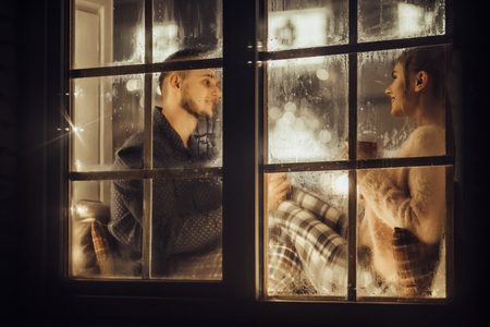 Young romantic couple is spending time together at home in pajamas.Sitting on window sill with garland in New Year Eve.Waiting for Christmas.Two lovers are hugging and kissing in Saint Valentine's Dayの写真素材