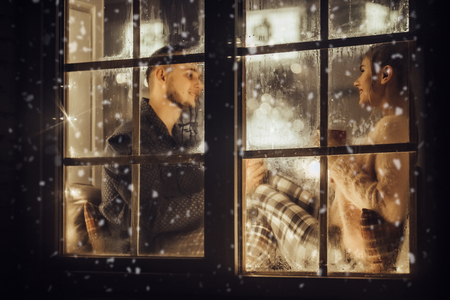 Young romantic couple is spending time together at home in pajamas.Sitting on window sill with garland in New Year Eve.Waiting for Christmas.Two lovers are hugging and kissing in Saint Valentine's Dayの写真素材