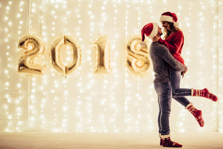 Young romantic couple in Santa Claus hats is waiting for Christmas at home together. Hugging and kissing in New Year Eve on the background of garlands and letters 2018.の写真素材