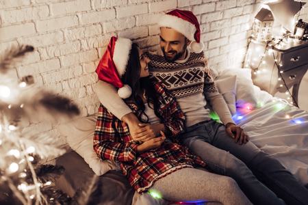 Merry Christmas and Happy New Year! Beautiful couple is lying in bed near Christmas tree in Santa Claus hats and hugging. Enjoying being together on the New Year's Eve.の写真素材