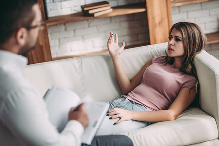 Young woman is lying on sofa during the psychotherapy session. Girl at a psychologist's reception. Experianced doctor is sitting nearby.の写真素材