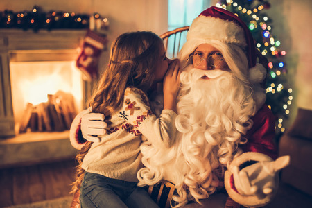 Little charming girl with happy Santa Claus in beautiful house decorated for Christmas. Child is telling secret wish on Santa's ear.の写真素材