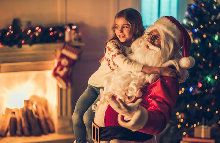Little charming girl with happy Santa Claus in beautiful house decorated for Christmas. Santa is holding child on hand and smiling.の写真素材