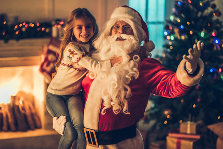 Little charming girl with happy Santa Claus in beautiful house decorated for Christmas. Santa is holding child on hand and smiling.の写真素材