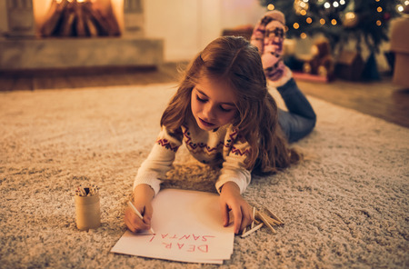 Little cute girl is waiting for Christmas at home. Charming kid is writing letter to Santa Claus in New Year Eve.の写真素材
