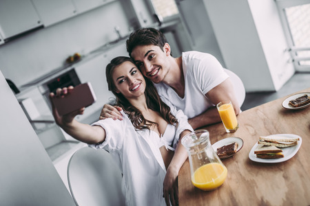 Good morning! Young romantic couple is having breakfast on modern light kitchen. Sexy woman in underwear and men's shirt and handsome man are sitting at the table and making selfie on smart phone. Healthy lifestyle concept.の写真素材