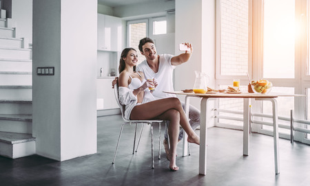 Good morning! Young romantic couple is having breakfast on modern light kitchen. Sexy woman in underwear and men's shirt and handsome man are sitting at the table and making selfie on smart phone. Healthy lifestyle concept.の写真素材