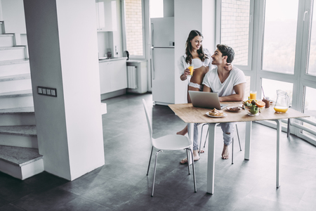 Good morning! Young romantic couple is having breakfast on modern light kitchen. Handsome man is sitting at the table with laptop while his sexy woman is standing nearby in underwear. Healthy lifestyle concept.の写真素材