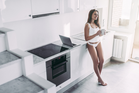 Young sexy woman in underwear is standing on light modern kitchen with laptop and cup of coffee in hands and smiling.の写真素材