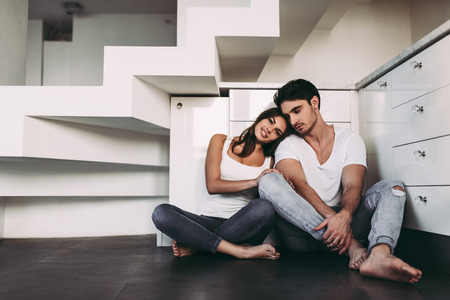 Romantic couple at home. Attractive young woman and handsome man are enjoying spending time together. Sitting on the floor in light modern kitchen.の写真素材