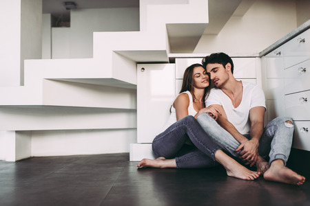 Romantic couple at home. Attractive young woman and handsome man are enjoying spending time together. Sitting on the floor in light modern kitchen.の写真素材