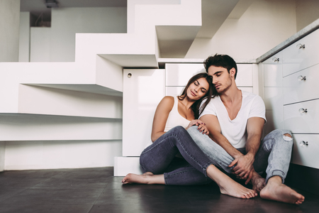 Romantic couple at home. Attractive young woman and handsome man are enjoying spending time together. Sitting on the floor in light modern kitchen.の写真素材