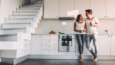 Full-length image of romantic couple at home. Attractive young woman and handsome man are enjoying spending time together while standing on light modern kitchen with cup of coffee in hands.の写真素材