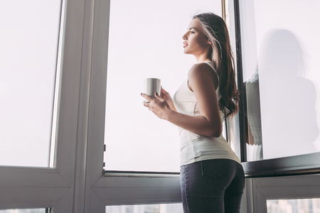 Attractive young woman is standing near open panoramic window with cup of coffee in hands.の写真素材