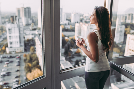 Attractive young woman is standing near open panoramic window with cup of coffee in hands. Beautiful view on city.の写真素材