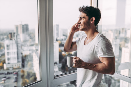 Handsome man is standing near open panoramic window with cup of coffee and smart phone in hands. Talking on mobile phone. Beautiful view on city.の写真素材