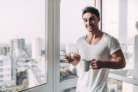 Handsome man is standing near open panoramic window with cup of coffee and smart phone in hands. Beautiful view on city.の写真素材
