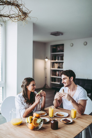 Good morning! Young romantic couple is having breakfast on modern light kitchen. Sexy woman in underwear and men's shirt and handsome man are sitting at the table, eating sandwiches and toasts, drinking orange juice. Healthy lifestyle concept.の写真素材