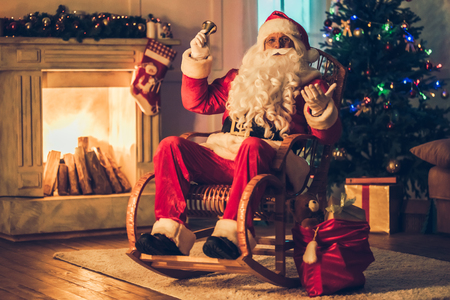 Happy Santa Claus in eyeglasses is preparing to Christmas in his residence, sitting in rocking chair, smiling and looking at camera.の写真素材
