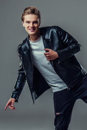Handsome young man isolated. Fashionable  man in leather jacket is standing on grey background, smiling and looking at camera.の写真素材