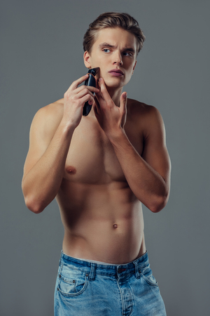 Handsome young man isolated. Portrait of shirtless muscular man is standing on grey background with trimmer in hand while shaving. Men care concept.の写真素材