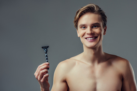 Handsome young man isolated. Portrait of shirtless muscular man is standing on grey background with razor in hand and smiling. Men care concept.の写真素材