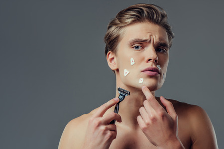 Handsome young man isolated. Portrait of shirtless muscular man is standing on grey background with razor in hand while shaving. Men care concept. Get hurt while shaving.の写真素材