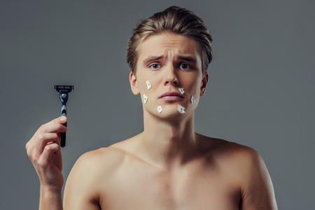 Handsome young man isolated. Portrait of shirtless muscular man is standing on grey background with razor in hand. Men care concept. Get hurt while shaving.の写真素材