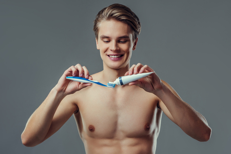 Handsome young man isolated. Portrait of shirtless muscular man is standing on grey background with toothbrush and toothpaste in hand. Men care concept.の写真素材