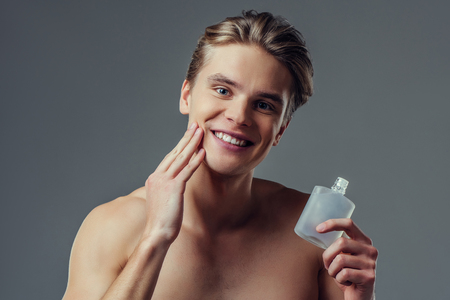 Handsome young man isolated. Portrait of shirtless muscular man is standing on grey background and using face aftershave lotion. Men care concept.の写真素材