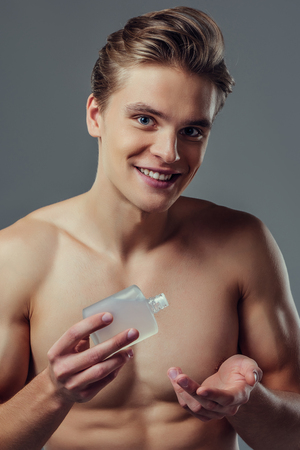 Handsome young man isolated. Portrait of shirtless muscular man is standing on grey background and using face aftershave lotion. Men care concept.の写真素材
