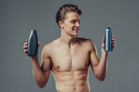 Handsome young man isolated. Portrait of shirtless muscular man is standing on grey background with shampoo in hands. Men care concept.の写真素材