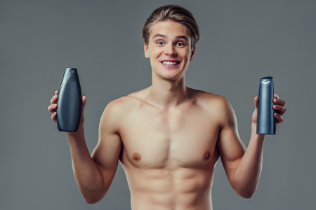 Handsome young man isolated. Portrait of shirtless muscular man is standing on grey background with shampoo in hands. Men care concept.の写真素材