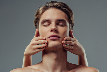 Handsome young man isolated. Portrait of shirtless muscular man is standing on grey background. Close-up of men's face while making face massage. Men care concept.の写真素材