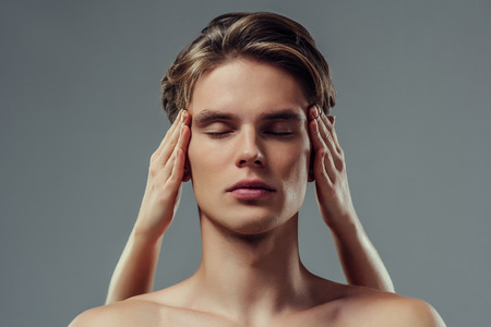 Handsome young man isolated. Portrait of shirtless muscular man is standing on grey background. Close-up of men's face while making face massage. Men care concept.の写真素材