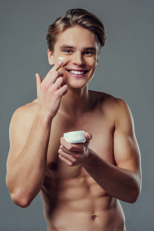 Handsome young man isolated. Portrait of shirtless muscular man is standing on grey background and using face cream, smiling and looking at camera. Men care concept.の写真素材