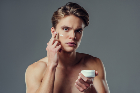 Handsome young man isolated. Portrait of shirtless muscular man is standing on grey background and using face cream. Men care concept.の写真素材