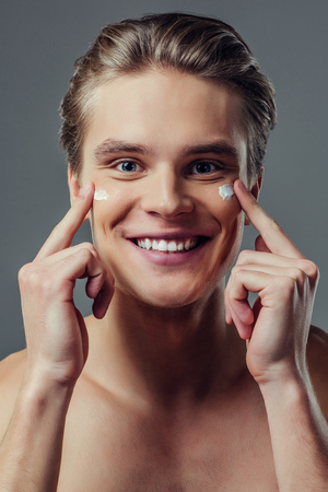 Handsome young man isolated. Portrait of shirtless muscular man is standing on grey background and using face cream, smiling and looking at camera. Men care concept.の写真素材