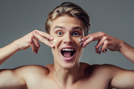 Handsome young man isolated. Portrait of shirtless muscular man is standing on grey background and using face cream, smiling and looking at camera. Men care concept.の写真素材