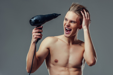 Handsome young man isolated. Portrait of shirtless muscular man is standing on grey background with hair dryer in hand. Men care concept.の写真素材