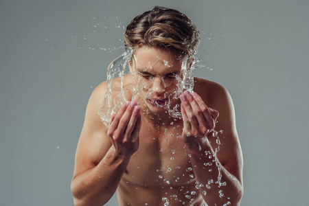 Handsome young man isolated. Portrait of shirtless muscular man is standing on grey background and washing oneself. Spraying water into face. Men care concept.の写真素材