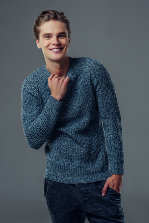 Handsome young man isolated. Fashionable  man in sweater is standing on grey background, smiling and looking at camera.の写真素材