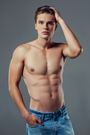 Handsome young man isolated. Shirtless muscular man in jeans is standing on grey background. Serious man looking at camera.の写真素材