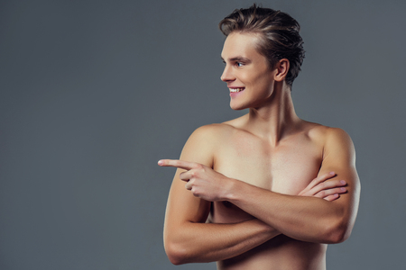 Handsome young man isolated. Portrait of shirtless muscular man is standing on grey background and pointing to the side. Showing product.の写真素材