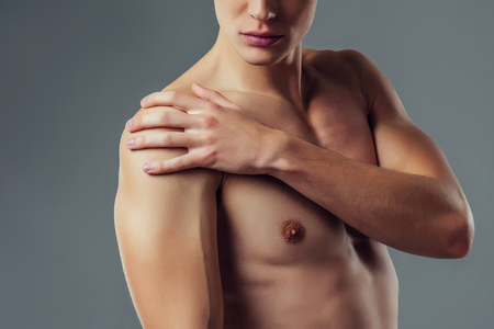 Handsome young man isolated. Cropped image of shirtless muscular man is standing on grey background. Close-up of man holding his shoulder. Experiencing shoulder pain.の写真素材