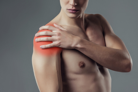 Handsome young man isolated. Cropped image of shirtless muscular man is standing on grey background. Close-up of man holding his shoulder. Experiencing shoulder pain.の写真素材