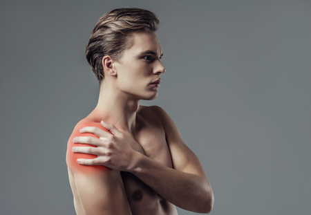Handsome young man isolated. Portrait of shirtless muscular man is standing on grey background. Man holding his shoulder. Experiencing shoulder pain.の写真素材