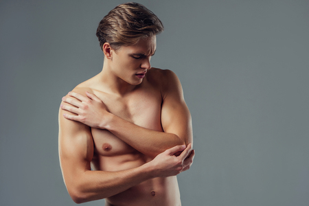 Handsome young man isolated. Portrait of shirtless muscular man is standing on grey background. Man holding his elbow. Experiencing elbow pain.の写真素材