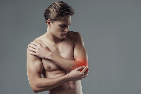 Handsome young man isolated. Portrait of shirtless muscular man is standing on grey background. Man holding his elbow. Experiencing elbow pain.の写真素材